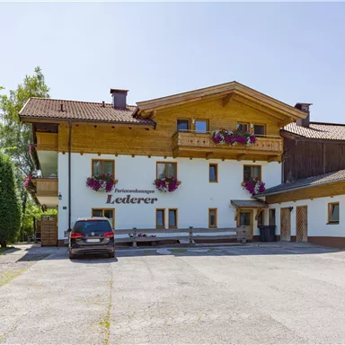 Ein Holzgebäude im bayerischen Stil mit Blumen auf den Fenstern. Vor dem Gebäude steht ein Auto auf einem befestigten Platz.