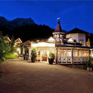 Ein charmantes Restaurant bei Nacht, umgeben von Bäumen und Bergen. Die beleuchtete Fassade sorgt für eine einladende Atmosphäre.