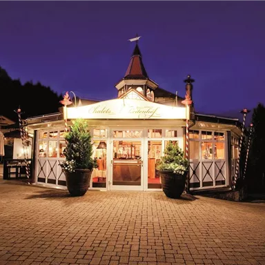 Ein hübsches Restaurant mit beleuchteter Fassade bei Dämmerung. Die Umgebung ist grün und einladend.