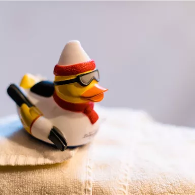 Eine lustige Gummiente mit Sonnenbrille und einer Mütze steht auf einem weißen Handtuch. Sie hat einen roten Schal um den Hals und sieht sportlich aus.