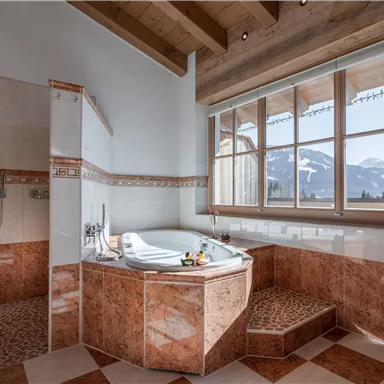 Ein elegantes Badezimmer mit einer großen Badewanne und einem Fenster, das den Blick auf die Berge freigibt. Die warmen Farbtöne und Holzdetails schaffen eine gemütliche Atmosphäre.
