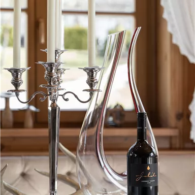 Eine elegante Tischdekoration mit einer Weinflasche, einem modernen Dekanter und silbernen Kerzenhaltern. Der Hintergrund ist hell und freundlich mit großen Fenstern.