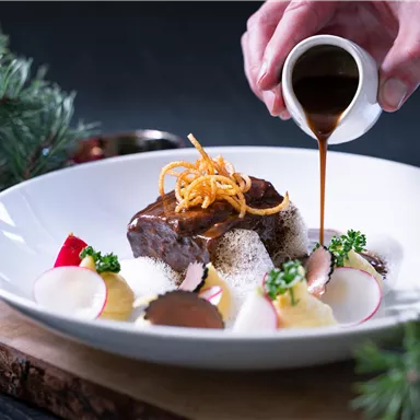 Ein gourmet Gericht auf einem weißen Teller, das mit einer Sauce übergossen wird. Es sind verschiedene Beilagen und eine ansprechende Präsentation zu sehen.