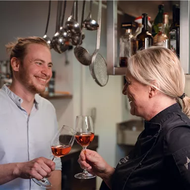 Zwei Personen stoßen mit Gläsern in einer Küche an. Beide lächeln und scheinen eine fröhliche Stimmung zu haben.