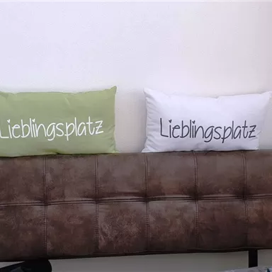 Zwei Kissen auf einer Couch mit der Aufschrift "Lieblingsplatz". Eines ist grün und das andere weiß.