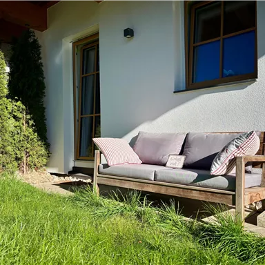A cozy bench is in the garden in front of a house. The grass is green and the sun is shining.