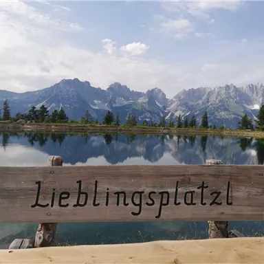 Ein ruhiger See mit Bergen im Hintergrund. Das Holzschild zeigt den Schriftzug "Lieblingsplatzl".