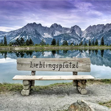 Eine Holzbank mit der Aufschrift „Lieblingsplatzl“ steht am Ufer eines klaren Sees. Im Hintergrund erheben sich majestätische Berge unter einem bewölkten Himmel.