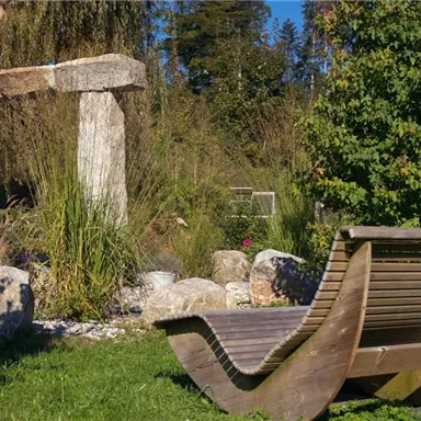 A tranquil garden landscape with a wooden lounge chair and a stone structure in the background. Surrounded by plants and stones, the place exudes a relaxed atmosphere.