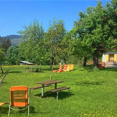 Eine grüne Wiese mit gemütlichen Liegestühlen und schattenspendenden Bäumen. Im Hintergrund ist ein Gebäude mit Balkonen und der Blick auf die Berge.