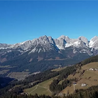 Eine beeindruckende Berglandschaft mit schneebedeckten Gipfeln und klarem blauen Himmel. Grüne Wiesen und kleine Dörfer liegen in der Täler.