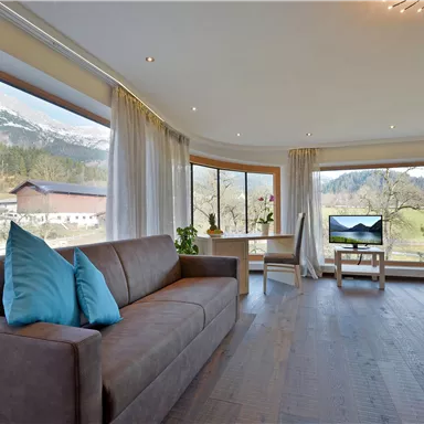 Ein modernes Wohnzimmer mit einem bequemen Sofa und bunten Kissen. Große Fenster bieten einen schönen Blick auf die umliegende Landschaft.