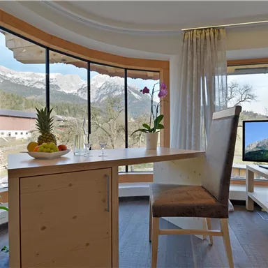 Ein modernes Wohnzimmer mit großen Fenstern und Blick auf Berge. Am Tisch stehen frisches Obst und eine Pflanze, neben einem Fernseher.