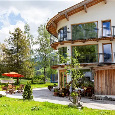 Eine moderne Villa mit Balkonen und einem schönen Garten. Im Vordergrund stehen Gartentische mit einem roten Sonnenschirm.