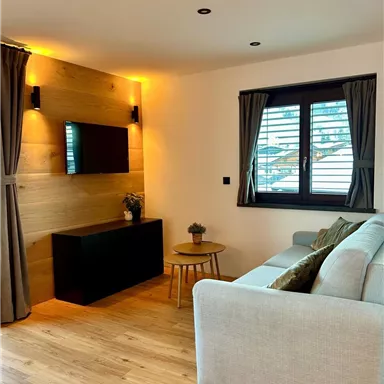 Ein modernes Wohnzimmer mit einem gemütlichen Sofa und einem Fernseher an der Wand. Große Fenster sorgen für viel Tageslicht und eine freundliche Atmosphäre.