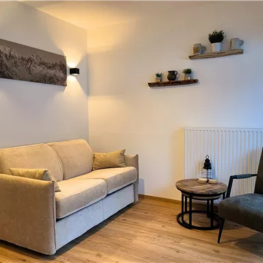 A cozy living room with a gray sofa and an armchair. Plant shelves and a picture hang on the wall.