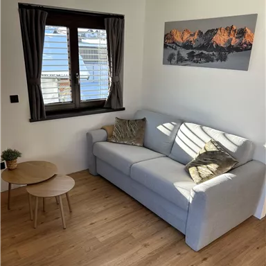 Ein gemütliches Wohnzimmer mit einem grauen Sofa und einem kleinen Tisch. Durch das Fenster scheint die Sonne auf den Holzfußboden.