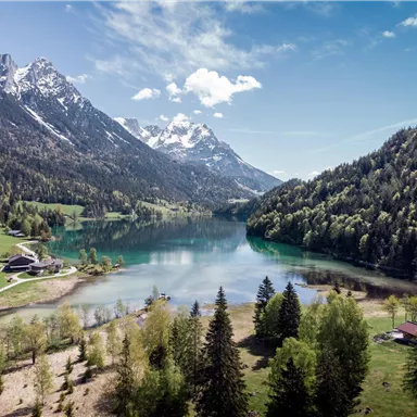 A picturesque landscape with a clear lake, surrounded by mountains and green meadows. The sun is shining and the sky is blue with some clouds.