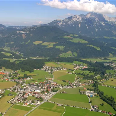 Eine malerische Landschaft mit sanften Hügeln und einem kleinen Dorf. Im Hintergrund sind majestätische Berge und ein blauer Himmel zu sehen.