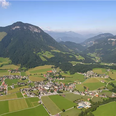 Eine malerische Landschaft mit sanften Hügeln und einem kleinen Dorf im Tal. Im Hintergrund erheben sich hohe Berge unter einem klaren Himmel.