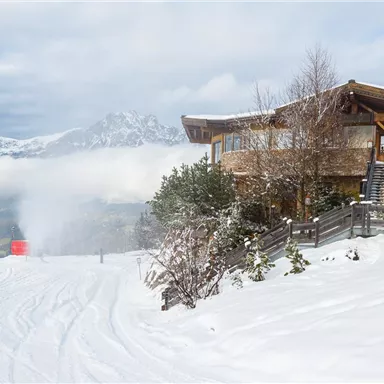 Eine verschneite Landschaft mit einem modernen Gebäude und schneebedeckten Bergen im Hintergrund. Am Fuße des Berges ist eine Skipiste zu sehen.