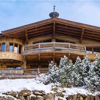 Ein gemütliches Holzhaus mit Balkon und großen Fenstern. Es liegt in einer verschneiten Landschaft mit Tannenbäumen.
