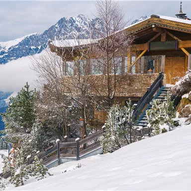 Ein gemütliches Holzhaus im Schnee, umgeben von Bäumen und Bergen. Der Himmel ist bewölkt und die Landschaft wirkt friedlich.
