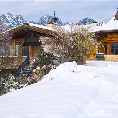 Ein gemütliches Holzhaus in einer verschneiten Landschaft. Im Hintergrund sind beeindruckende Berge zu sehen.