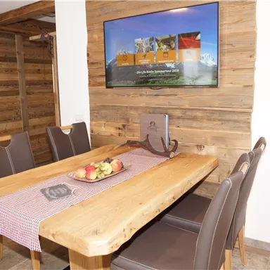 Ein gemütlicher Essbereich mit einem großen Holztisch und modernen Stühlen. An der Wand hängt ein Fernseher, und auf dem Tisch liegt eine Schüssel mit Äpfeln.