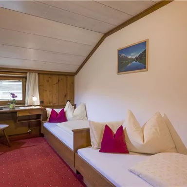 Ein gemütlicher Raum mit zwei Betten, einem kleinen Tisch und einem Fenster. An der Wand hängt ein Bild von einer Berglandschaft.