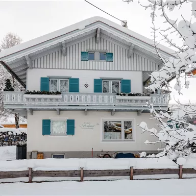 A charming house in the snow with blue shutters. The surroundings are wintry and peaceful, surrounded by snow-covered trees.