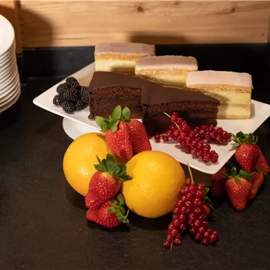 Ein Teller mit verschiedenen Kuchenstücken, darunter Schokoladenkuchen und andere Süßigkeiten. Daneben befindet sich eine Auswahl an frischen Früchten wie Erdbeeren, Johannisbeeren und Orangen.