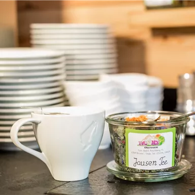 Eine Tasse mit Tee steht neben einem Glas mit Tee-Zutaten. Im Hintergrund sind stapelweise weiße Teller zu sehen.