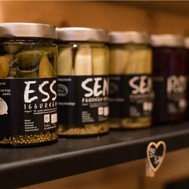 Eine Reihe von eingelegten Gurken und Oliven in klaren Gläsern mit ansprechenden Etiketten. Die Gläser stehen auf einem Regal aus Holz.