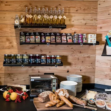 Ein rustikales Frühstücksbufett mit frischen Brötchen, Käse und Früchten. Regale sind mit Marmeladen und Ölen gefüllt, umgeben von einer Holzverkleidung.