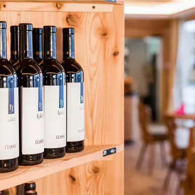 Fünf Flaschen Rotwein stehen auf einem Holzregal. Im Hintergrund erkennt man einen gemütlichen Essbereich.