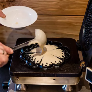 Eine Person gießt Teig in eine spezielle Form auf einer Heizplatte. Der Teig wird gleichmäßig verteilt, um ein rundes Gebäck herzustellen.