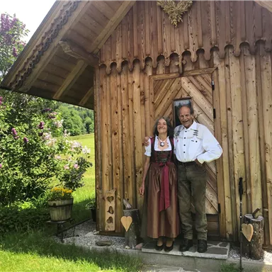 Ein Paar steht vor einer schönen Holzblockhütte. Die Frau trägt ein traditionelles Dirndl, während der Mann in einer Tracht gekleidet ist.