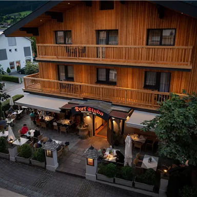 A cozy restaurant with a terrace and wooden facade. The surroundings are green and inviting, perfect for a relaxing evening.