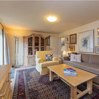 Ein gemütliches Wohnzimmer mit einem beigen Sofa und einem gelben Sessel. Es gibt einen Holztisch, Bücherregale und dekorative Bilder an den Wänden.