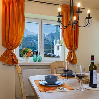 Ein gemütlicher Essbereich mit einem Holztisch, Geschirr und Wein. Der Blick durch das Fenster zeigt die Berge und eine Kirche im Hintergrund.
