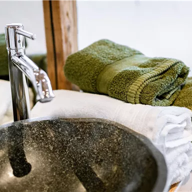 Ein modernes Waschbecken aus Stein mit glänzenden Wasserhähnen. Darauf liegen stapelweise grüne und weiße Handtücher.