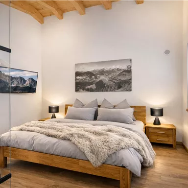 Ein modernes Schlafzimmer mit einem großen Bett, weichen Decken und Holzmöbeln. Große Fenster sorgen für viel Tageslicht und einen angenehmen Ambiente.