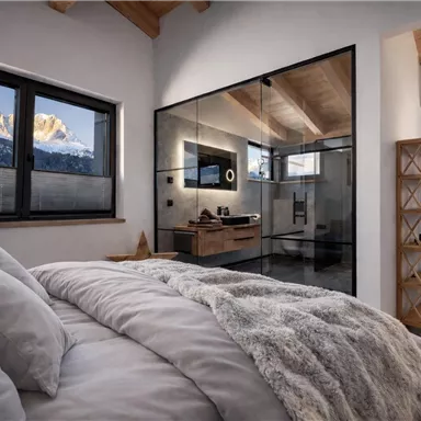 Ein modernes Schlafzimmer mit einem komfortablen Bett und einem Blick auf schneebedeckte Berge. Das Bad ist durch eine Glaswand getrennt und strahlt Eleganz aus.