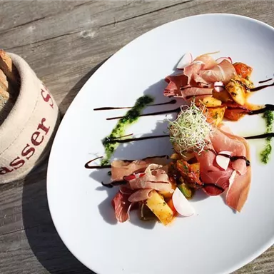 Ein kunstvoll angerichteter Teller mit verschiedenen Schinkensorten und Gemüse, garniert mit Sprossen. Neben dem Teller liegt eine Portion Brot in einem Korb.