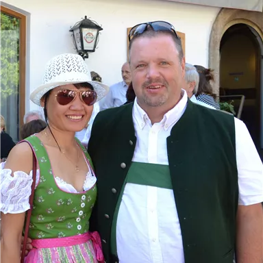 Ein Paar in traditioneller bayerischer Kleidung steht zusammen. Die Frau trägt ein Dirndl, während der Mann in einer Tracht gekleidet ist.
