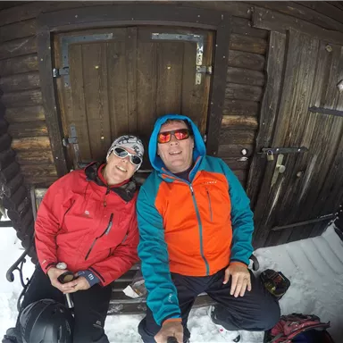 Ein Paar sitzt in einer gemütlichen Hütte im Schnee. Beide tragen sportliche Kleidung und Sonnenbrillen, während sie lächeln.