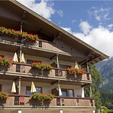 Ein traditionelles alpenländisches Gebäude mit blühenden Balkonen. Im Hintergrund ist ein beeindruckender Berg und ein blauer Himmel zu sehen.