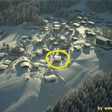 Eine schneebedeckte Landschaft mit mehreren Häusern in einer hügeligen Umgebung. In der Mitte ist ein Gebäude hervorgehoben, umgeben von weißem Schnee.