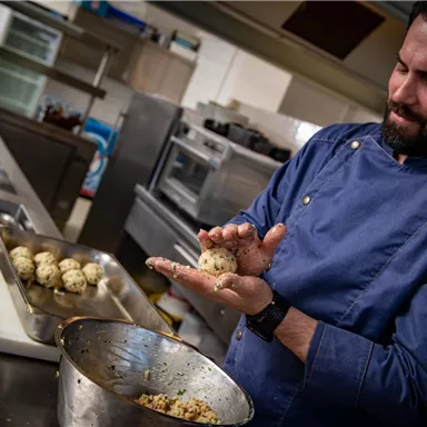 Ein Koch formt Teigbälle in einer modernen Küche. Im Hintergrund sind weitere Zutaten und Utensilien zu sehen.